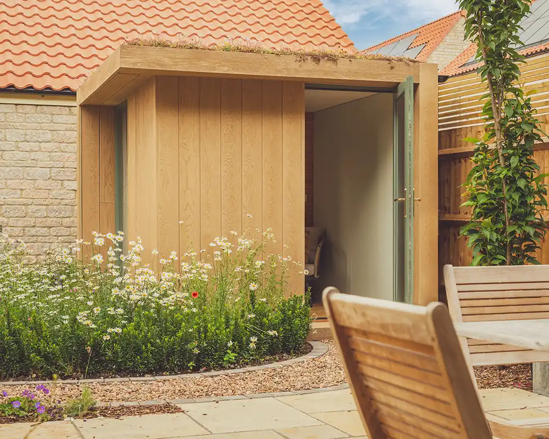 Golden oak office pod in the corner of a an english garden surrounded by flowers and a gravel path